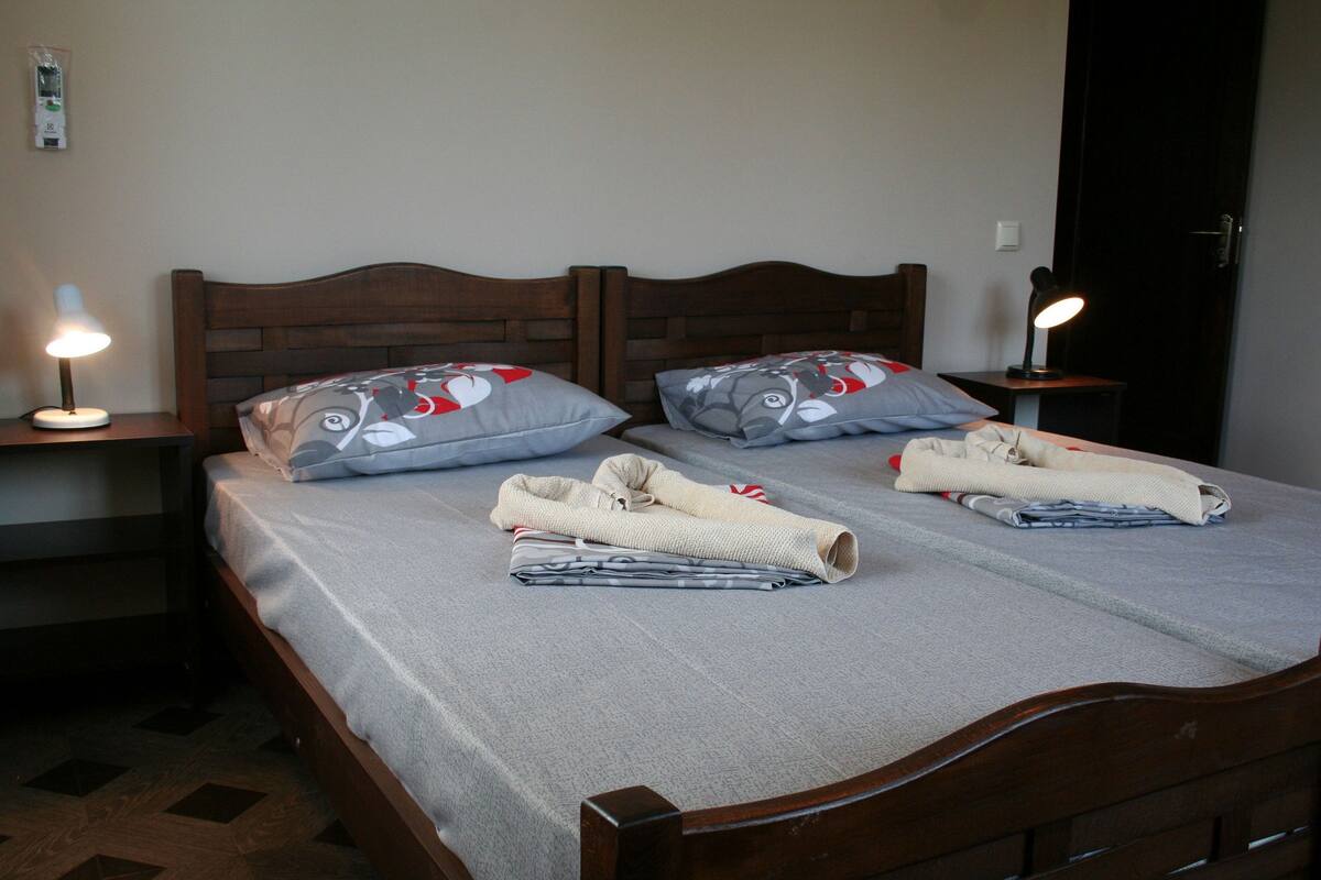 A double bed with a grey bedspread, two pillows with a red and grey pattern, and two folded towels. There is a bedside table with a lamp on the left side.