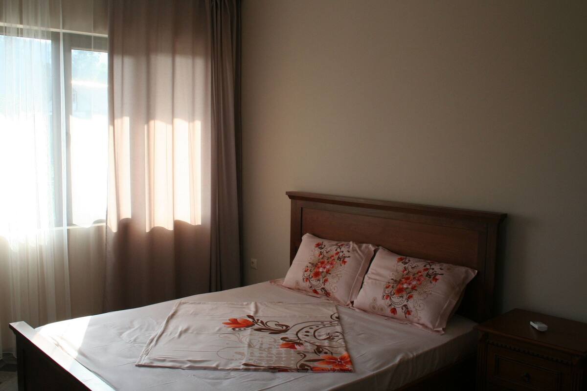 A neatly made bed with floral bedding, a wooden headboard, and a nightstand with a remote control.