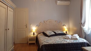 A bedroom with a wooden headboard, a chandelier, bedside lamps, a bed with a floral duvet, and a window with curtains. - A L'ombre Du Clocher (Calenzana)