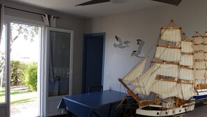 A room with a large window, a ceiling fan, a table with a blue tablecloth, and a model ship on a stand. - A L'ombre Du Clocher (Calenzana)