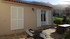 A building with a stone patio, two white shutters, a small outdoor seating area, and a view of mountains in the background. - A L'ombre Du Clocher (Calenzana)