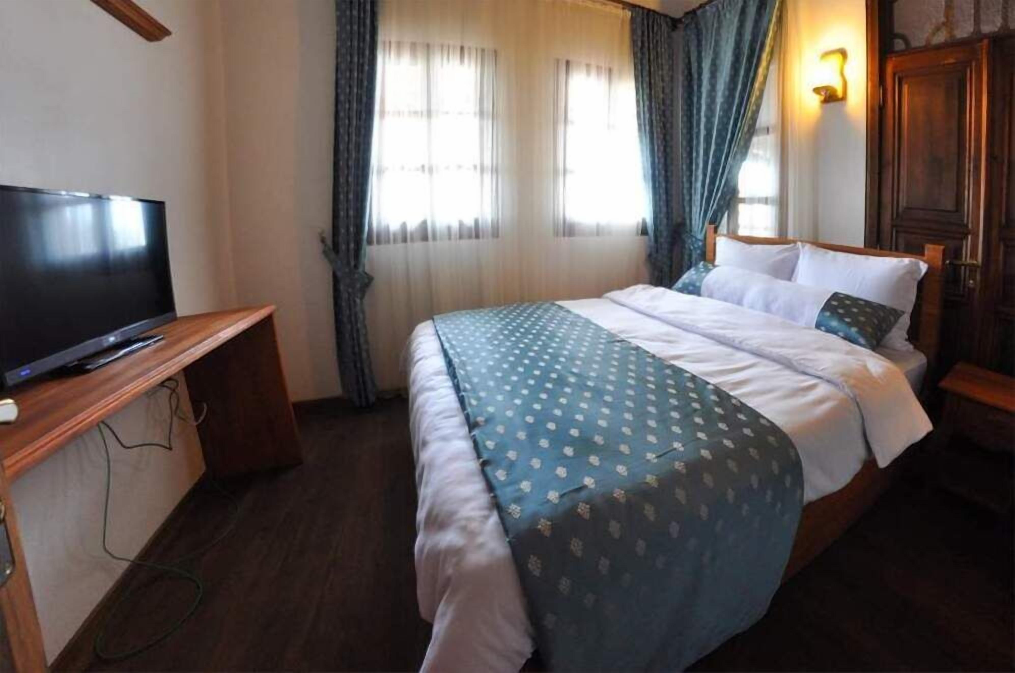 A hotel room with a bed, a TV on a wooden stand, a window with curtains, and a wall-mounted light.