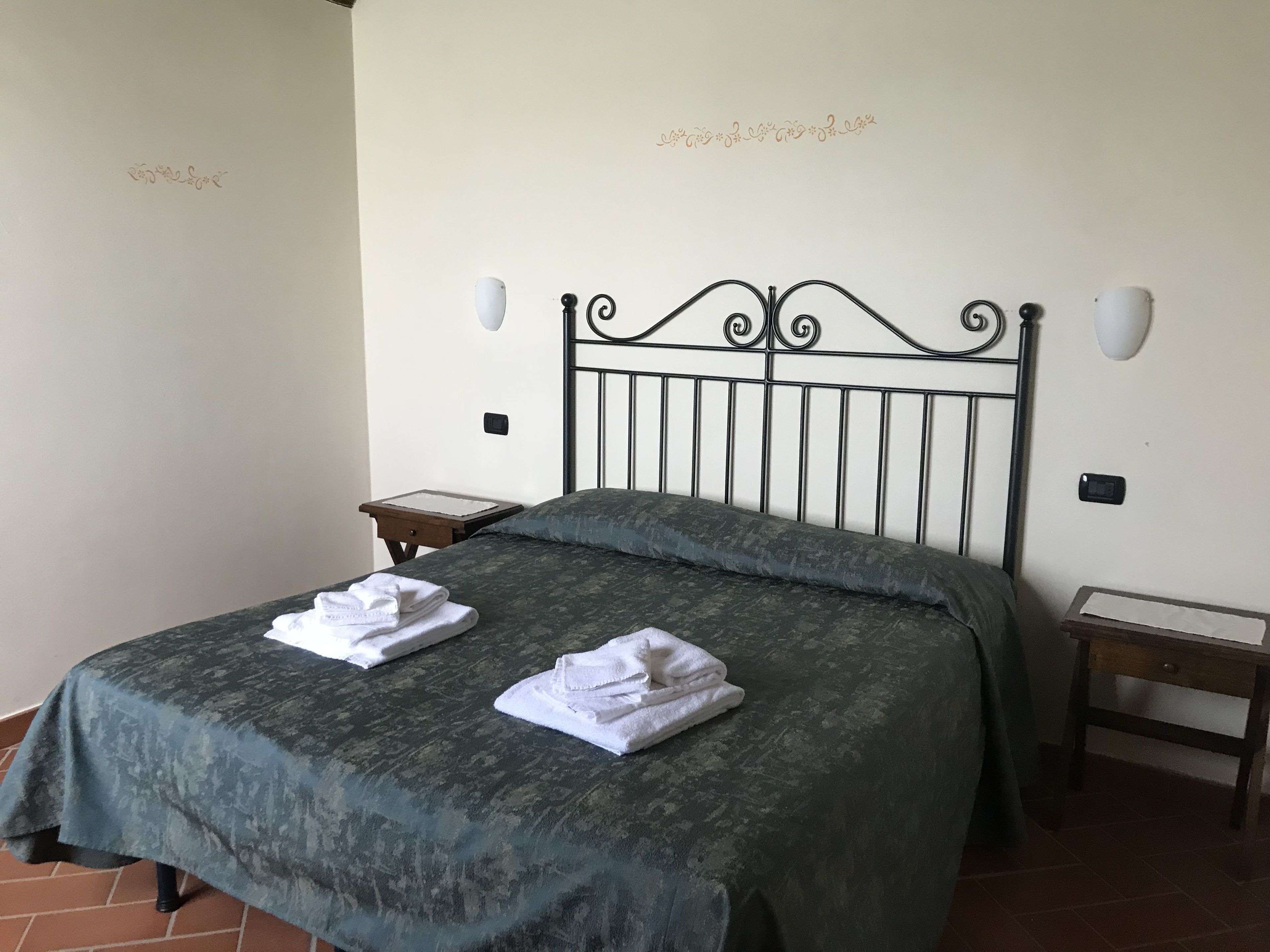 Un letto rifatto con una coperta verde e due asciugamani bianchi piegati. Due comodini, uno con una lampada e l'altro con un piccolo oggetto sopra. Pareti lisce con scritte decorative.