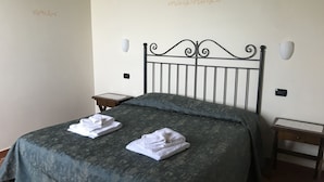 Una cama bien hecha con una colcha verde y dos toallas blancas dobladas. Hay dos mesitas de noche, una con una lámpara y la otra con un objeto pequeño. Las paredes son lisas con escritura decorativa.