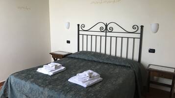 Un letto rifatto con una coperta verde e due asciugamani bianchi piegati. Due comodini, uno con una lampada e l'altro con un piccolo oggetto sopra. Pareti lisce con scritte decorative.