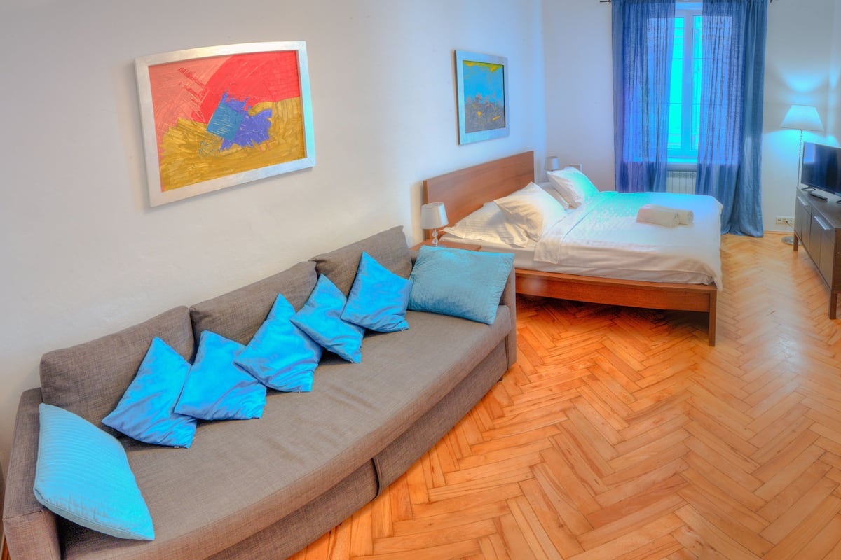 A modern hotel room with a wooden herringbone floor, a sofa with blue cushions, a bed with white linens, and two framed artworks on the walls.