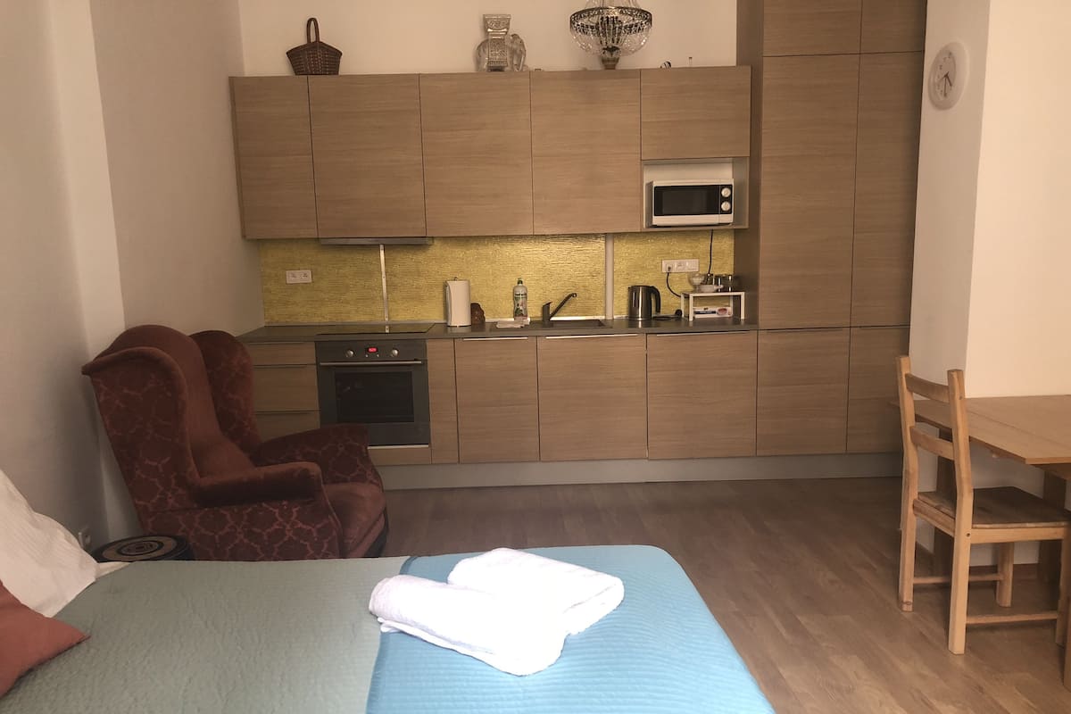 A compact room with a bed, a chair, a small table, a kitchenette, and a wooden floor.