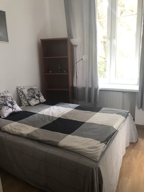 A neatly made bed with a checkered bedspread, a bedside lamp, and a bookshelf with a few items on the lower shelf. - Rais Serviced Apartments (Warsaw)