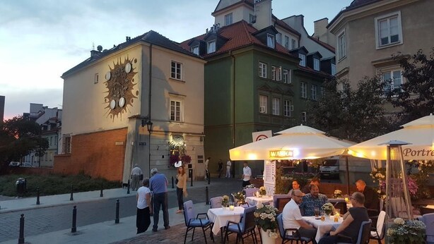 An outdoor dining area with tables and chairs, a clock on a building, and a sign that reads "Warka". - Rais Serviced Apartments (Warsaw)
