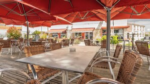 Un área de comedor al aire libre con sillas y mesas de mimbre, sombrillas rojas y un edificio de piedra al fondo.