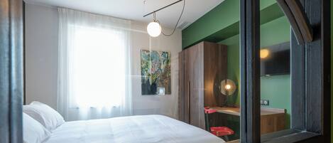 Habitación de hotel con cama, ventana con cortinas, un cuadro en la pared y vista a la cocina a través de la ventana.