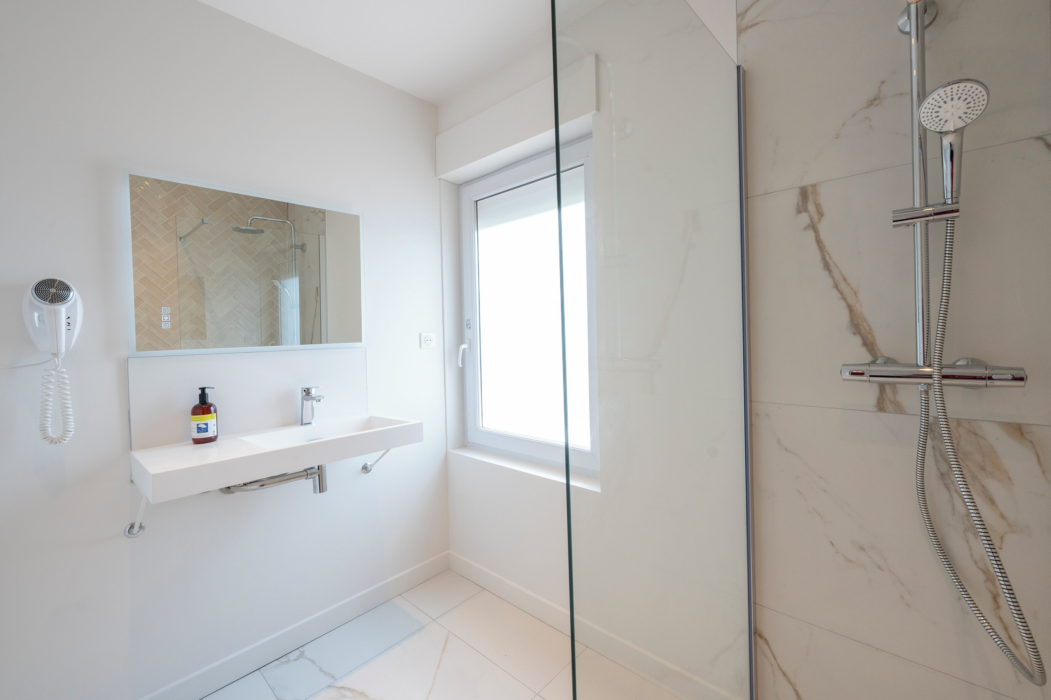 A modern bathroom with a glass shower enclosure, a white sink with a mirror above, and a bottle of hand soap on the sink.