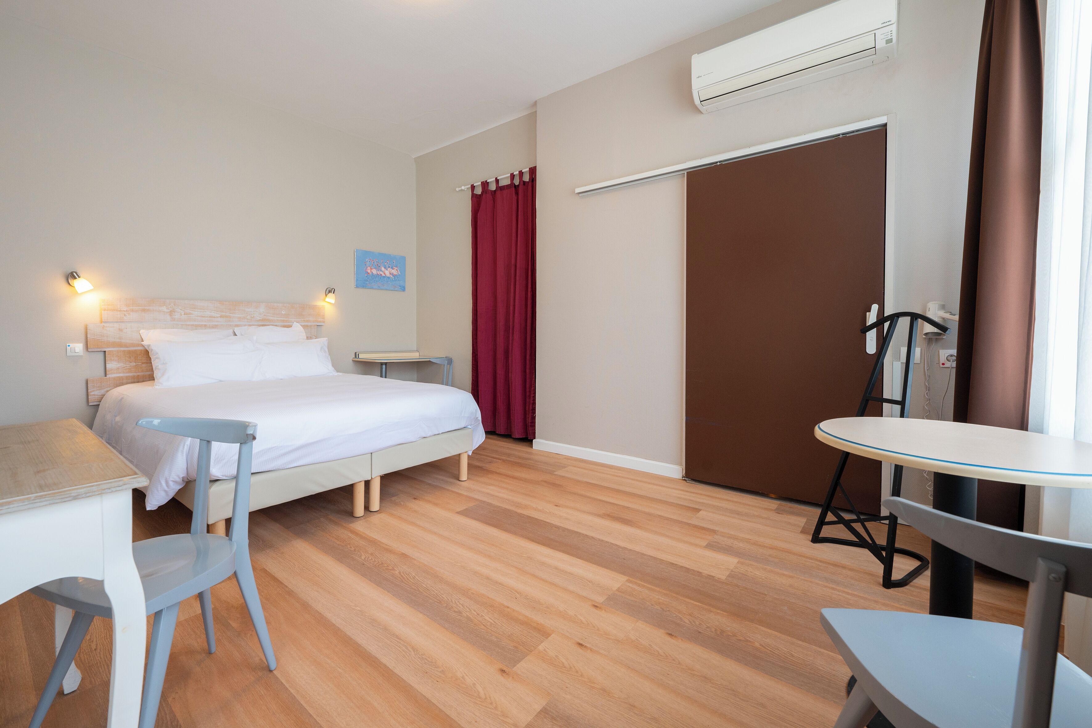 A hotel room with a bed, a desk, a chair, a table, a wardrobe, and an air conditioning unit.