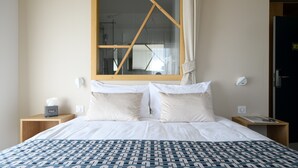 Habitación de hotel con una cama grande, dos almohadas, una mesita de noche con caja de pañuelos y una lámpara, y un ventanal con marco de madera.