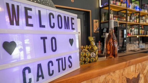 Una zona de bar con un letrero que dice 'WELCOME TO CACTUS', mostrador de madera y estantes con botellas.