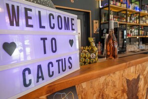 A bar area with a 'WELCOME TO CACTUS' sign, wooden counter, and shelves with bottles. - Hôtel Les Pins - Oléron  (Saint-Trojan-les-Bains)