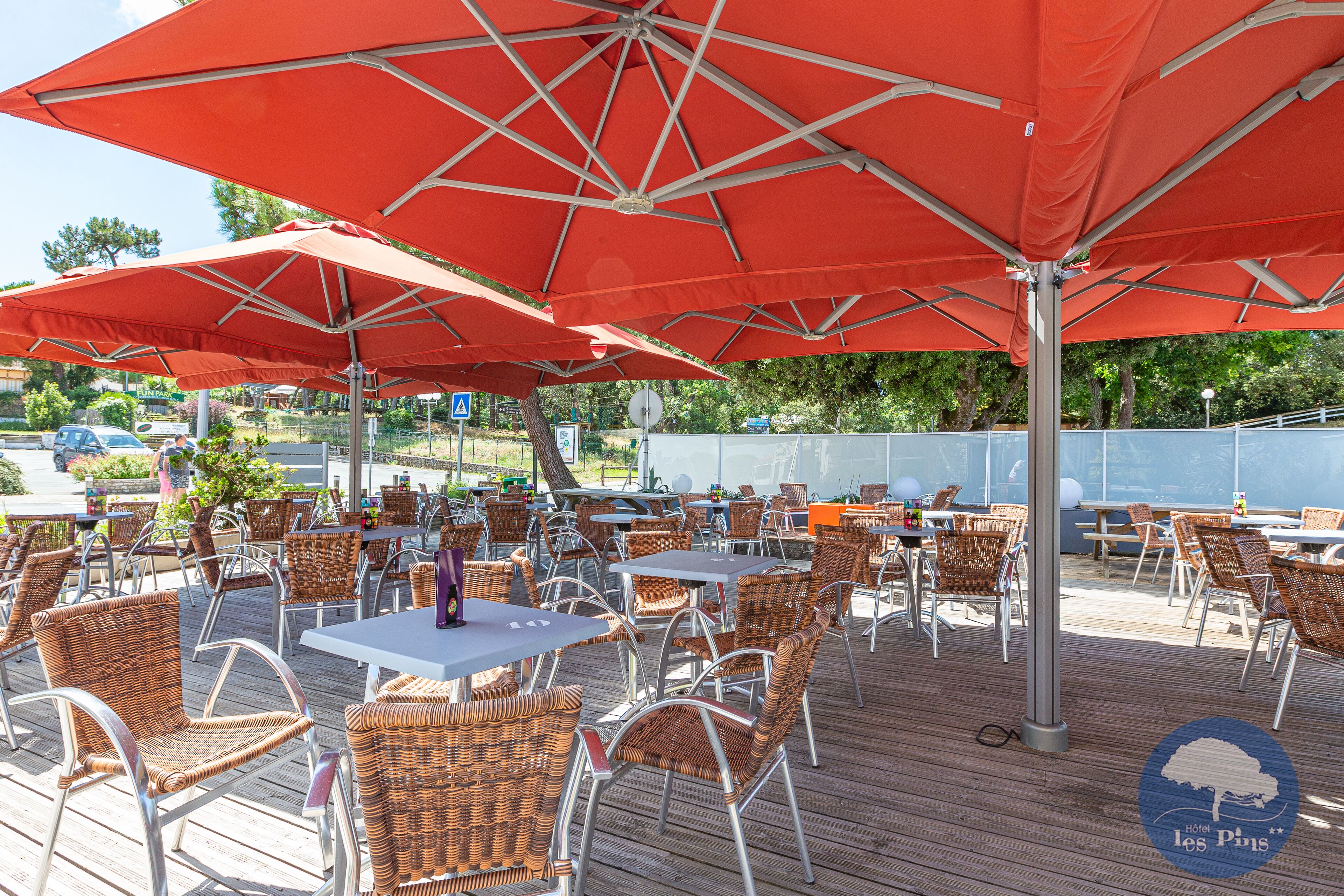 Een terras buiten met rieten stoelen en tafels, rode parasols en het logo van 'Hotel les Pins'.