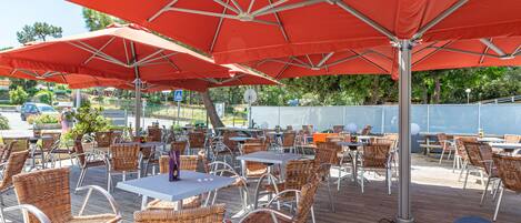 Een terras buiten met rieten stoelen en tafels, rode parasols en het logo van 'Hotel les Pins'.