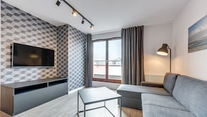 Een moderne woonkamer met een grijze bank, een flatscreen-tv aan een muur met een geometrisch patroon, een klein tafeltje en een vloerlamp.
