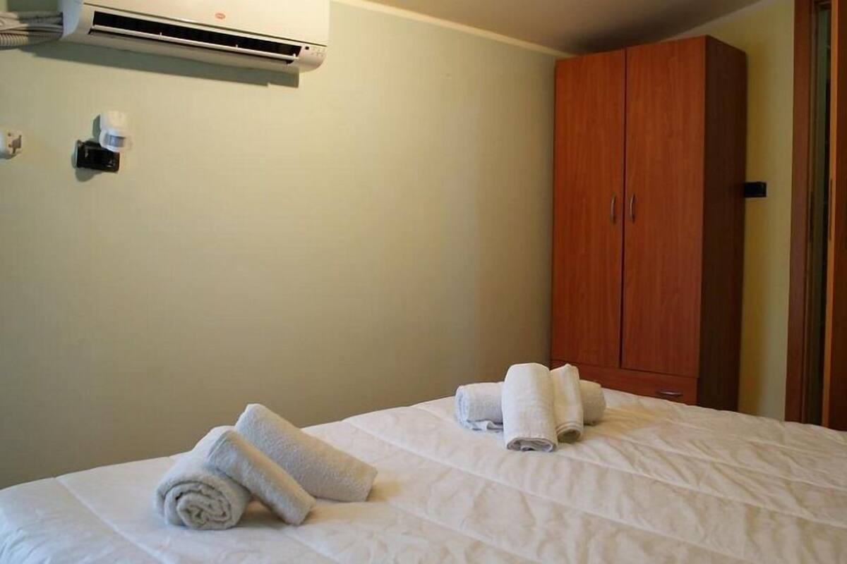 A hotel room with a bed, two rolled towels, an air conditioning unit, and a wooden wardrobe.