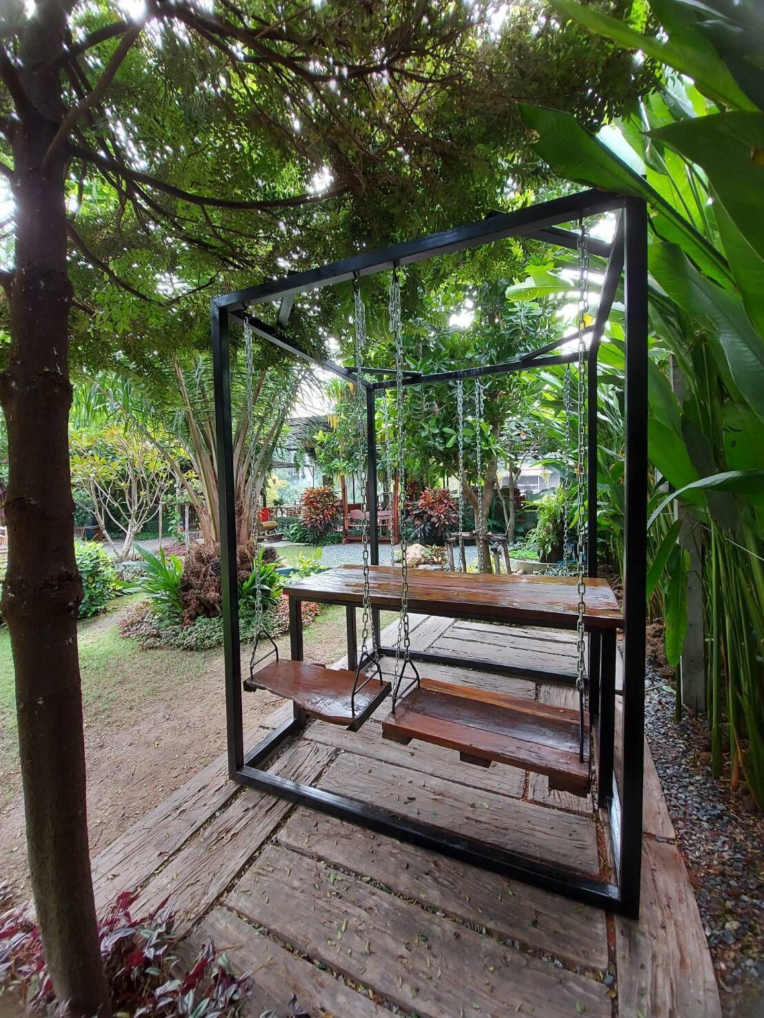 Un columpio de madera con estructura metálica, rodeado de abundante vegetación y un jardín.