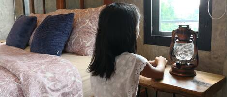 Un niño sentado a una mesa de madera con un farol, en una habitación con una cama y almohadones estampados.