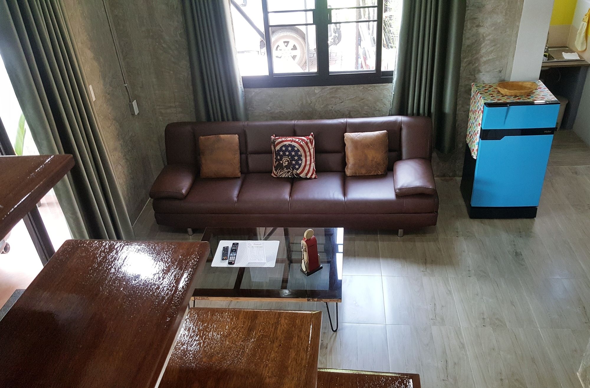 A brown leather sofa with three cushions, a glass coffee table, and a blue cabinet in a room with concrete walls and a window.