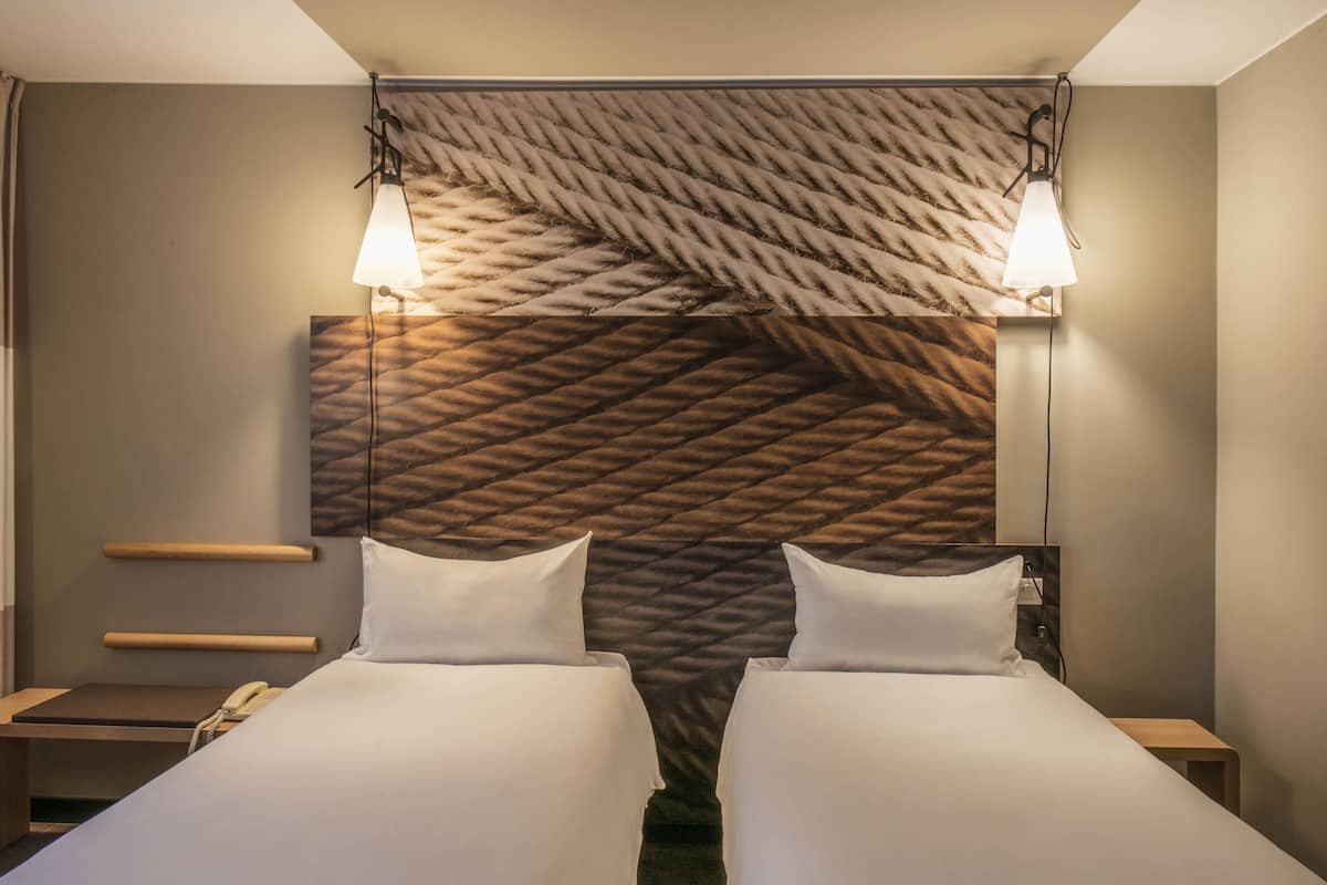 A hotel room with two beds, a headboard with a textured pattern, bedside lamps, a telephone, and a small wooden table.