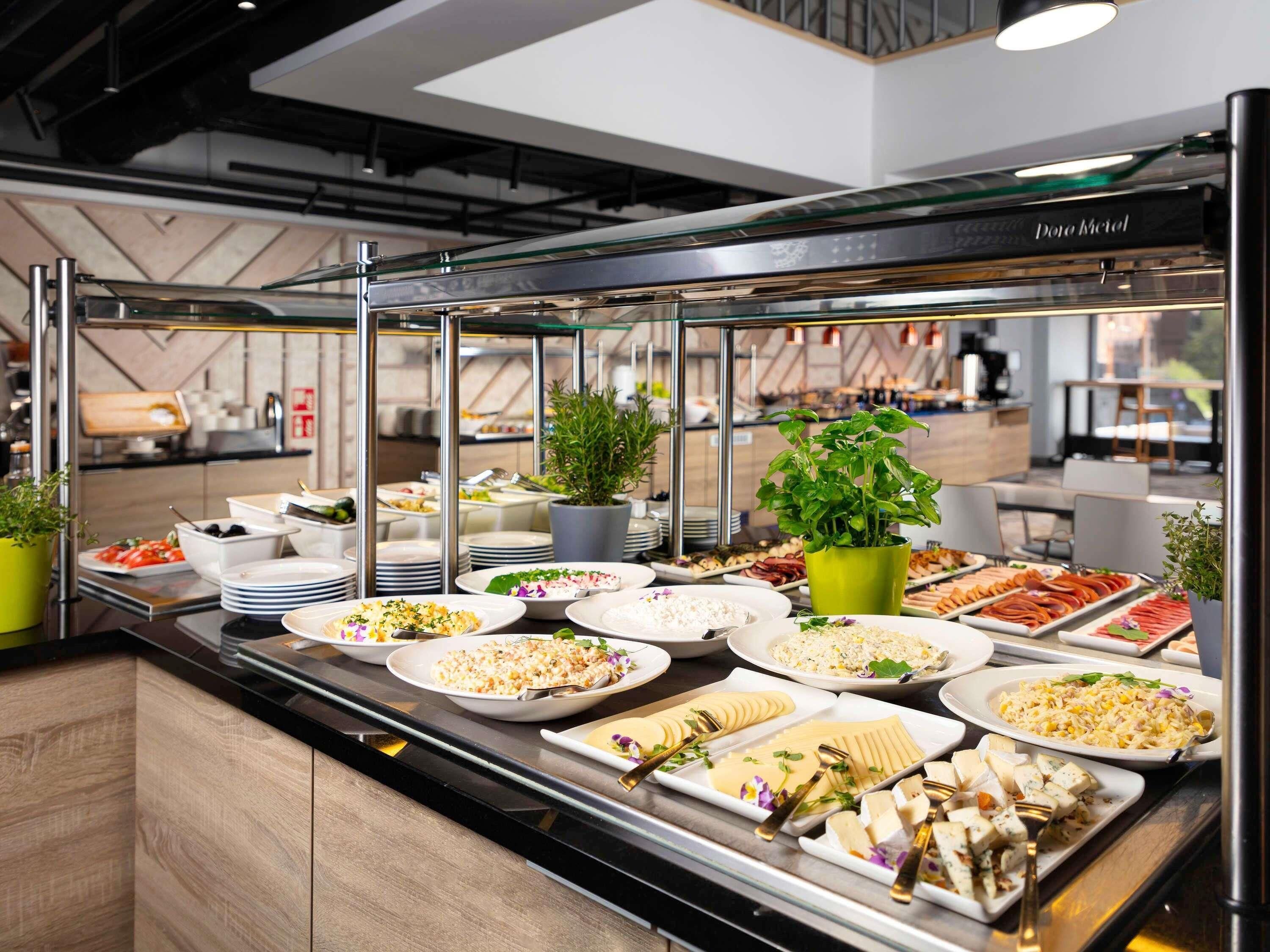Ein reichhaltiges Buffet mit verschiedenen Speisen, darunter Salate, Fleisch und Käse, präsentiert auf einer Theke mit einer Glasschutzwand.