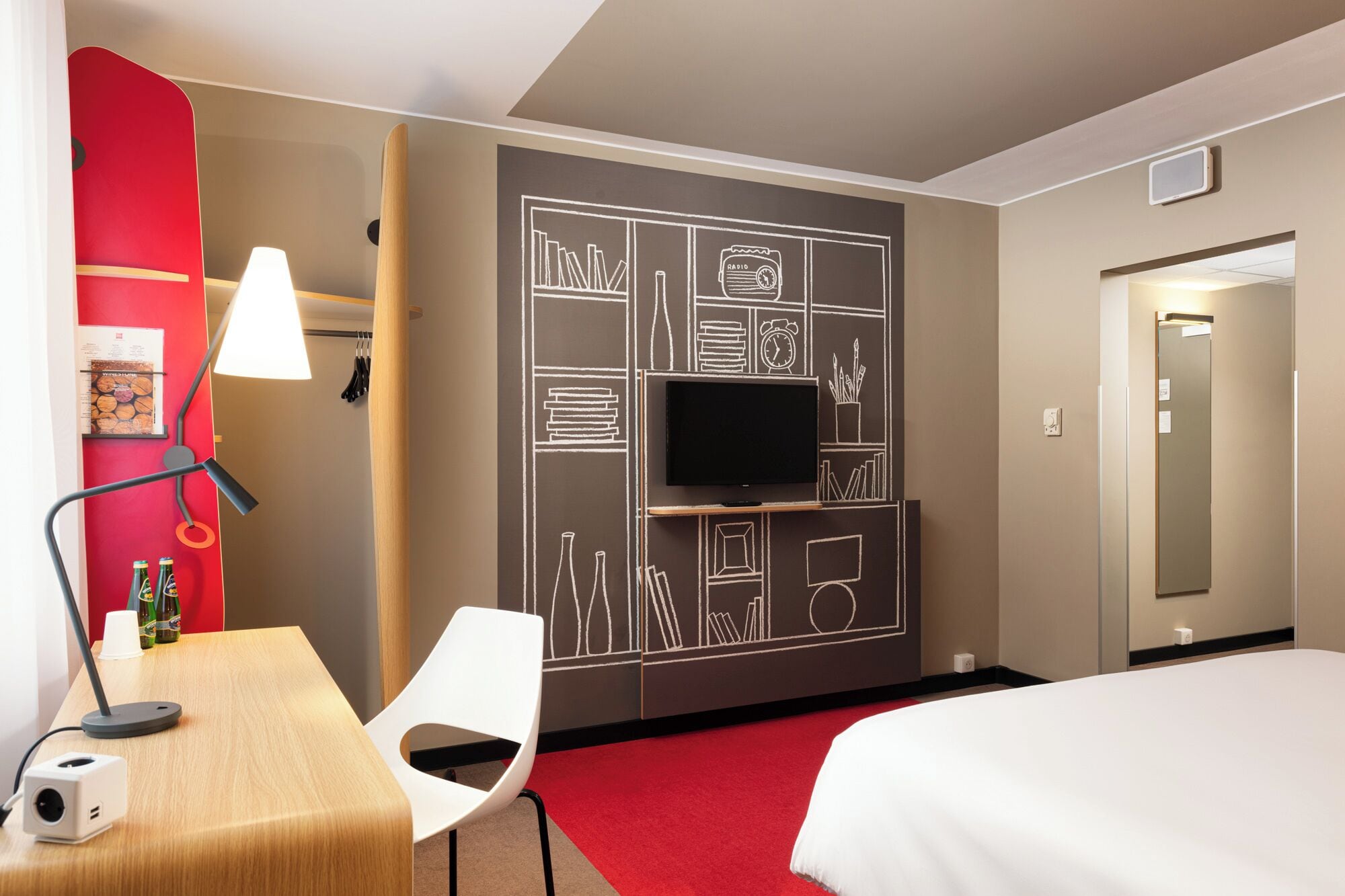 A hotel room with a bed, a desk with a lamp, a chair, a TV mounted on a wall with a chalkboard design, and a red accent wall.