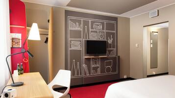 A hotel room with a bed, a desk with a lamp, a chair, a TV mounted on a wall with a chalkboard design, and a red accent wall.