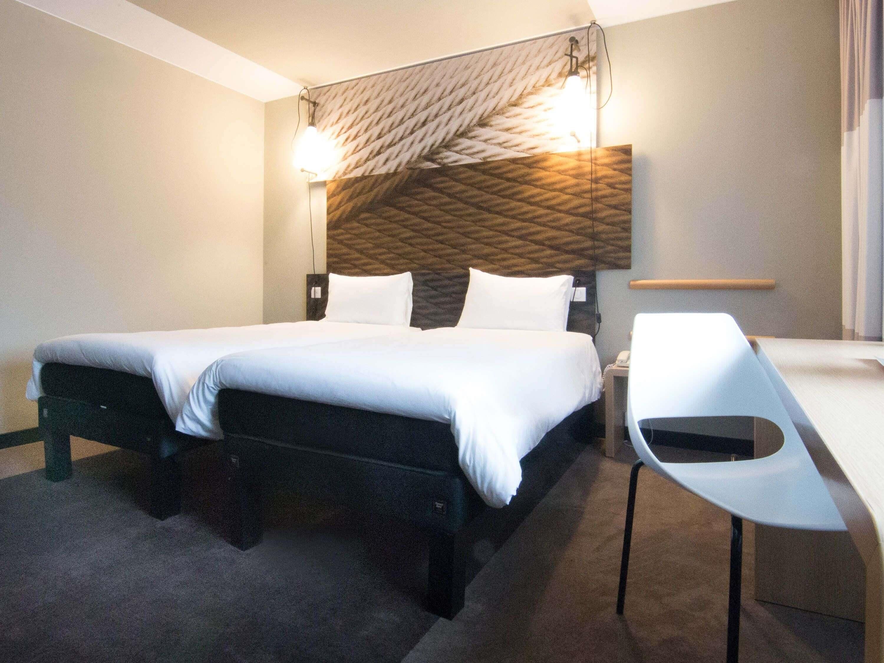 A hotel room with a bed, bedside tables, a desk, and a wall-mounted light fixture.