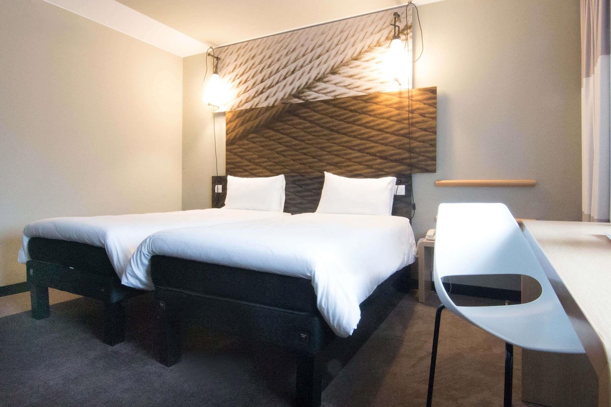Una habitación de hotel con cama, mesitas de noche, un escritorio y una lámpara de pared.