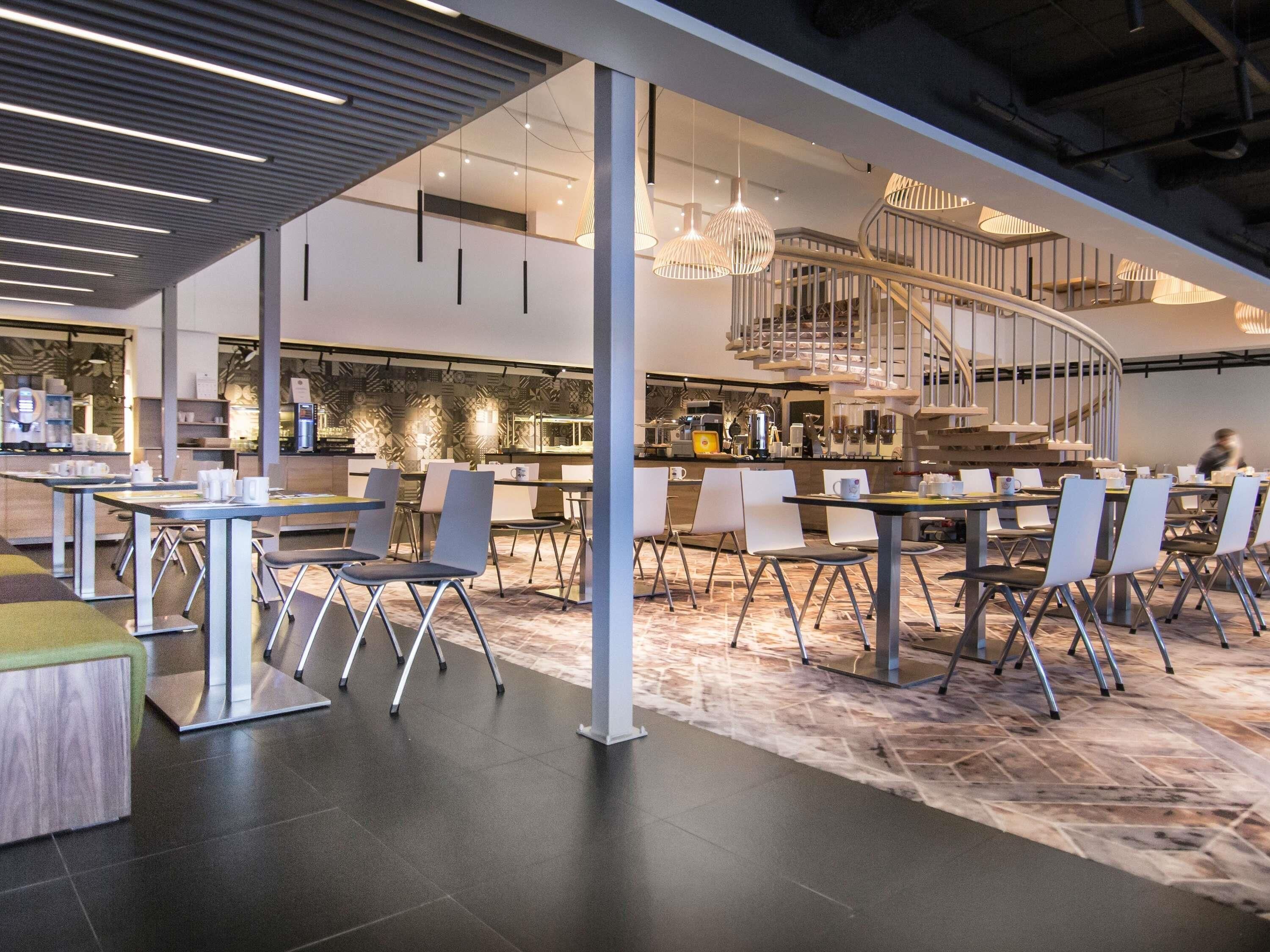 Un café moderno con una escalera central, mesas, sillas y una zona de cocina.