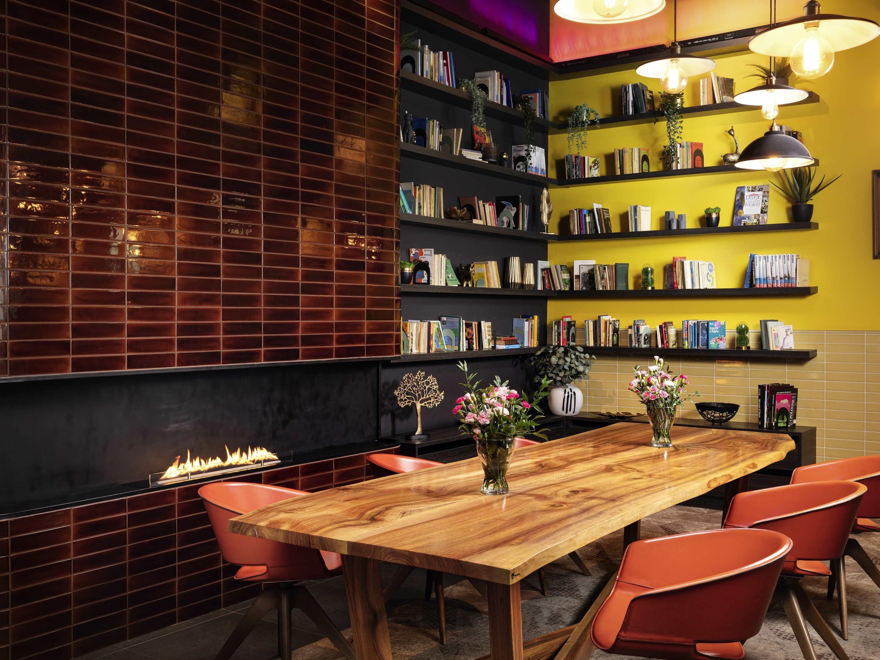 A modern interior with a wooden table, red chairs, bookshelves, and a fireplace.