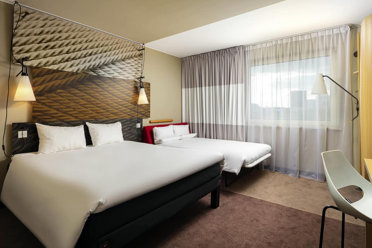 A hotel room with a large bed, a smaller bed, a desk, a chair, and a window with curtains.
