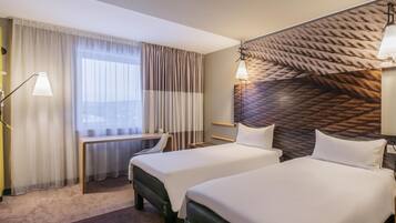 A hotel room with two beds, a desk, a chair, a lamp, and a view of the city.