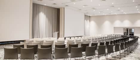 Un salón de conferencias con filas de sillas, una cortina grande y una pantalla de proyector.