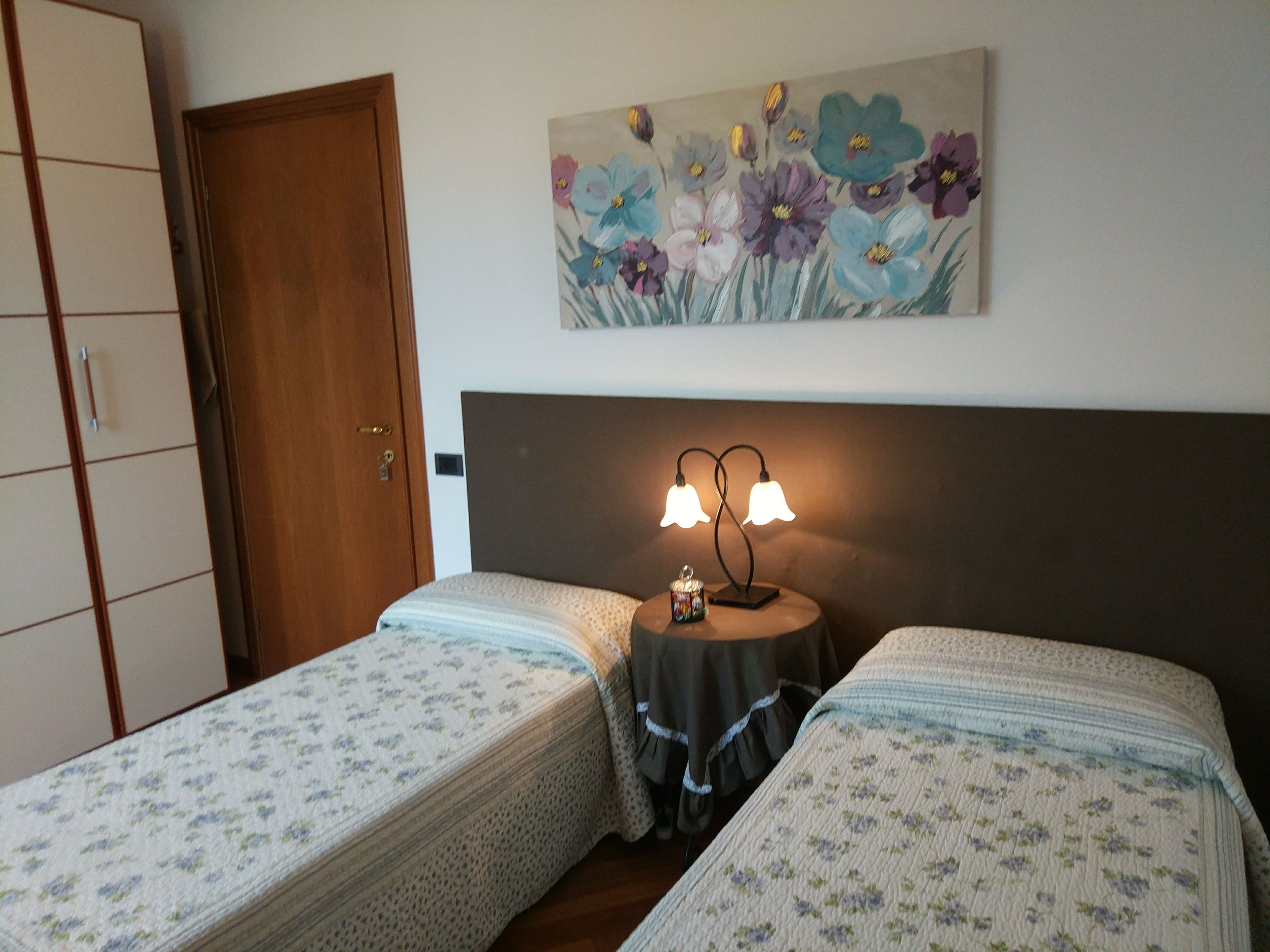 Un dormitorio con dos camas individuales, un armario de madera, un cuadro de flores en la pared y una mesita de noche con una lámpara.