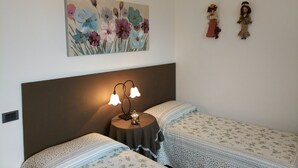 A bedroom with two beds, a nightstand with a lamp, a floral painting, and two figurines on the wall. - Marinella B (Cornale e Bastida)
