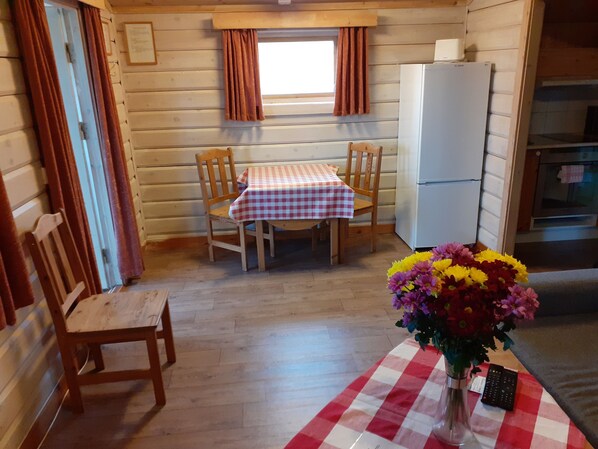 Una habitación acogedora con una mesa y sillas de madera, un ventanal con cortinas, un refrigerador y un florero con flores.