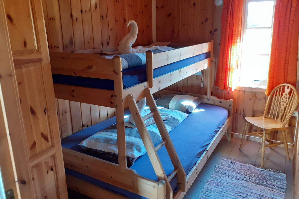 Habitación con literas, paredes de madera, una ventana con cortinas naranjas y una silla.