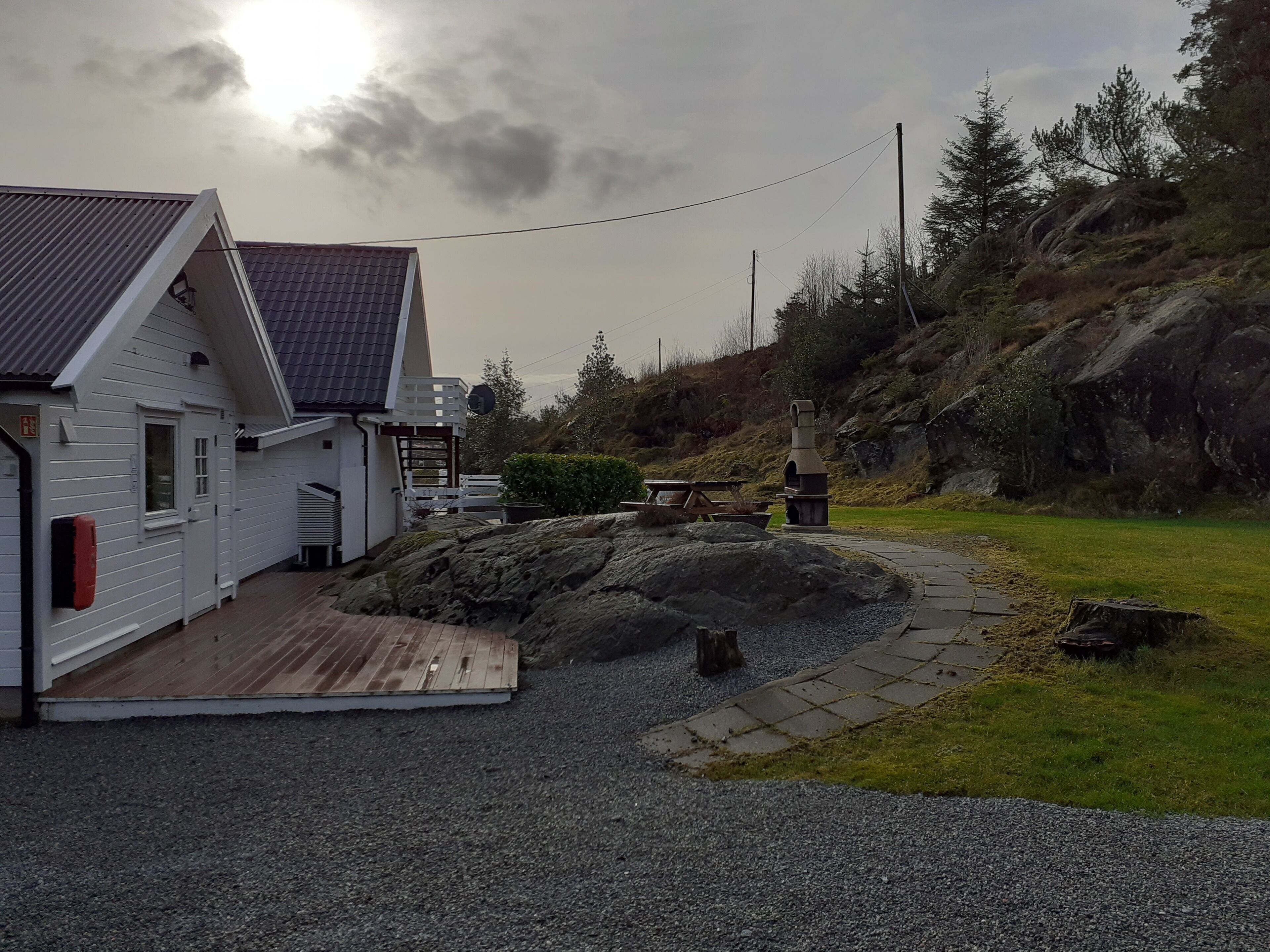 A white building with a sloped roof, a wooden deck, and a stone pathway leading to a picnic table and outdoor grill.