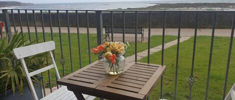 Un balcon avec une table en bois et deux chaises blanches, un vase rempli de fleurs et une vue sur la mer.