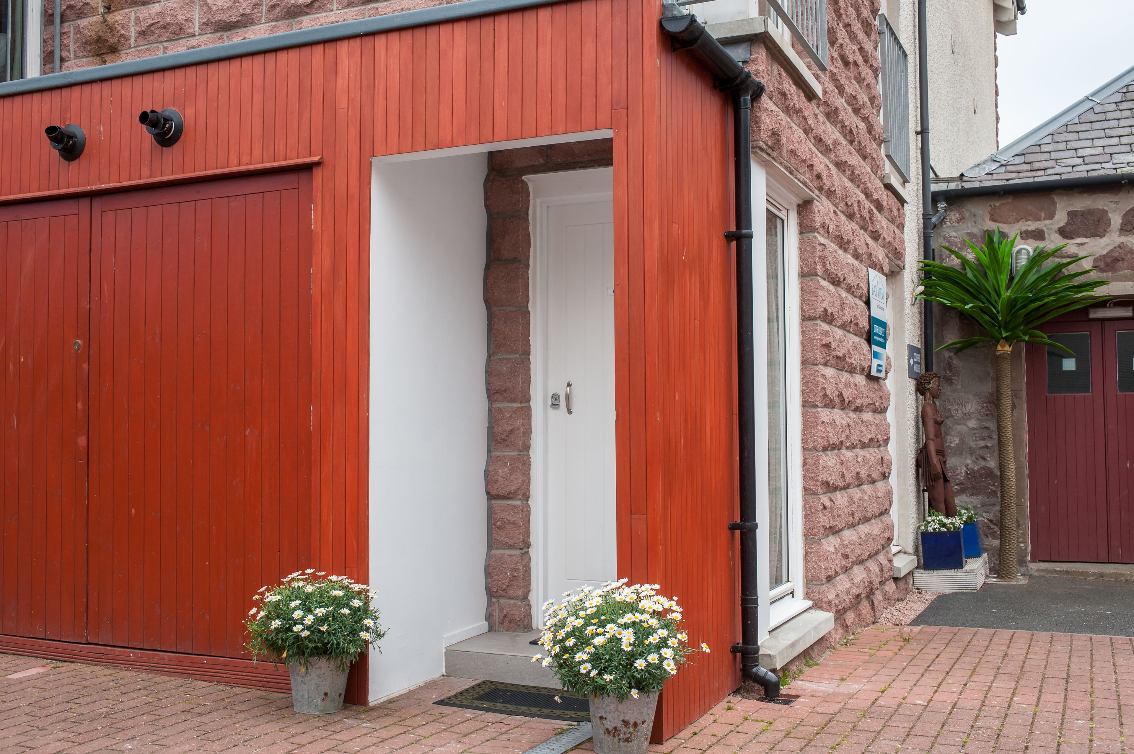 A red building with a white door, two potted plants, and a stone wall.