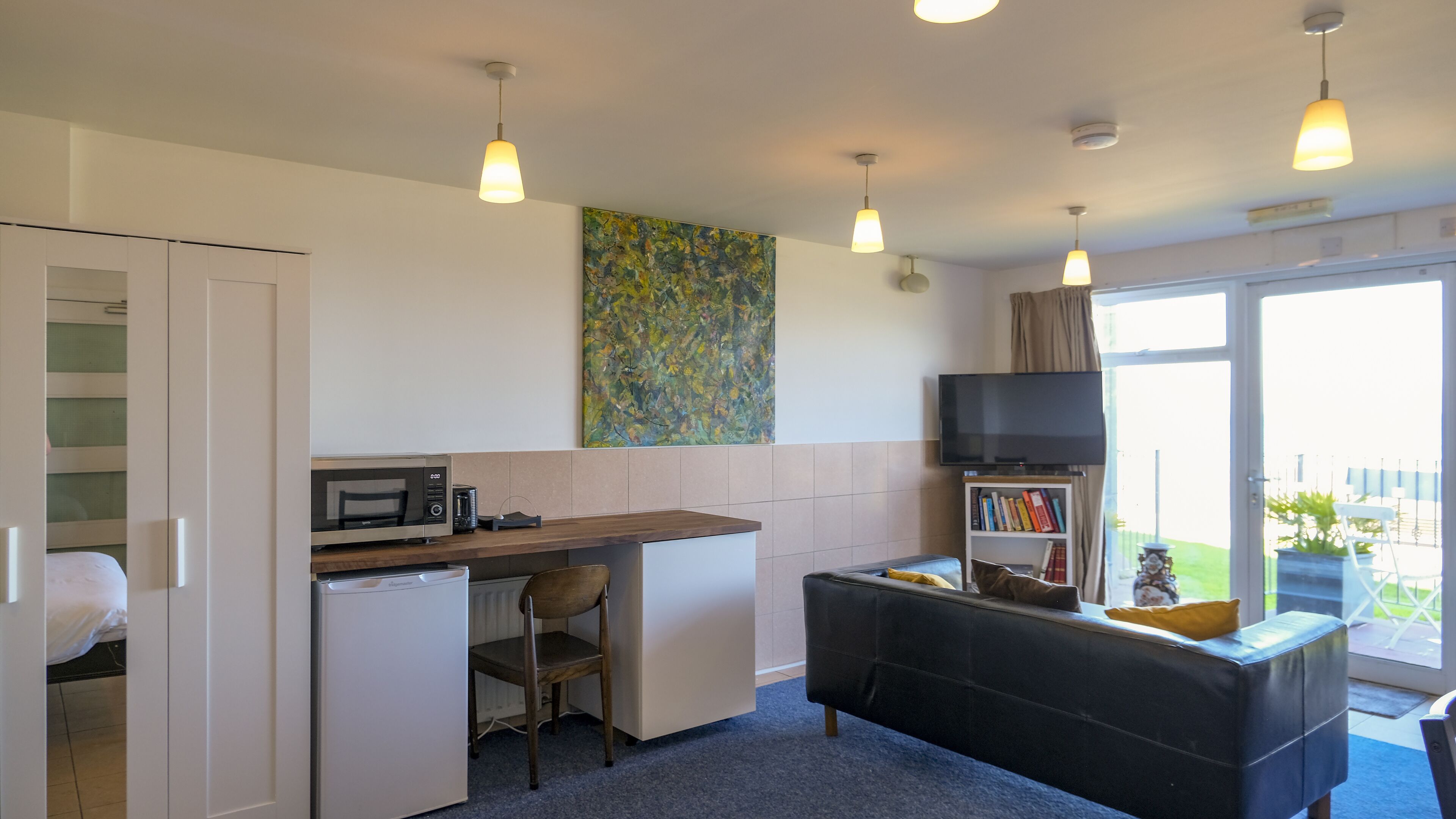 A compact kitchen with a microwave, a small refrigerator, and a dining area with a chair. There is a bookshelf with books and a flat-screen TV mounted on the wall. The room has a large window providing a view of the outdoors.