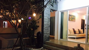 A modern house with a stone exterior, a glass door leading to a living room with a sofa and cushions, and a small garden area with pebbles and a tree. - Casa D Pool Huahin 1 (Hua Hin)