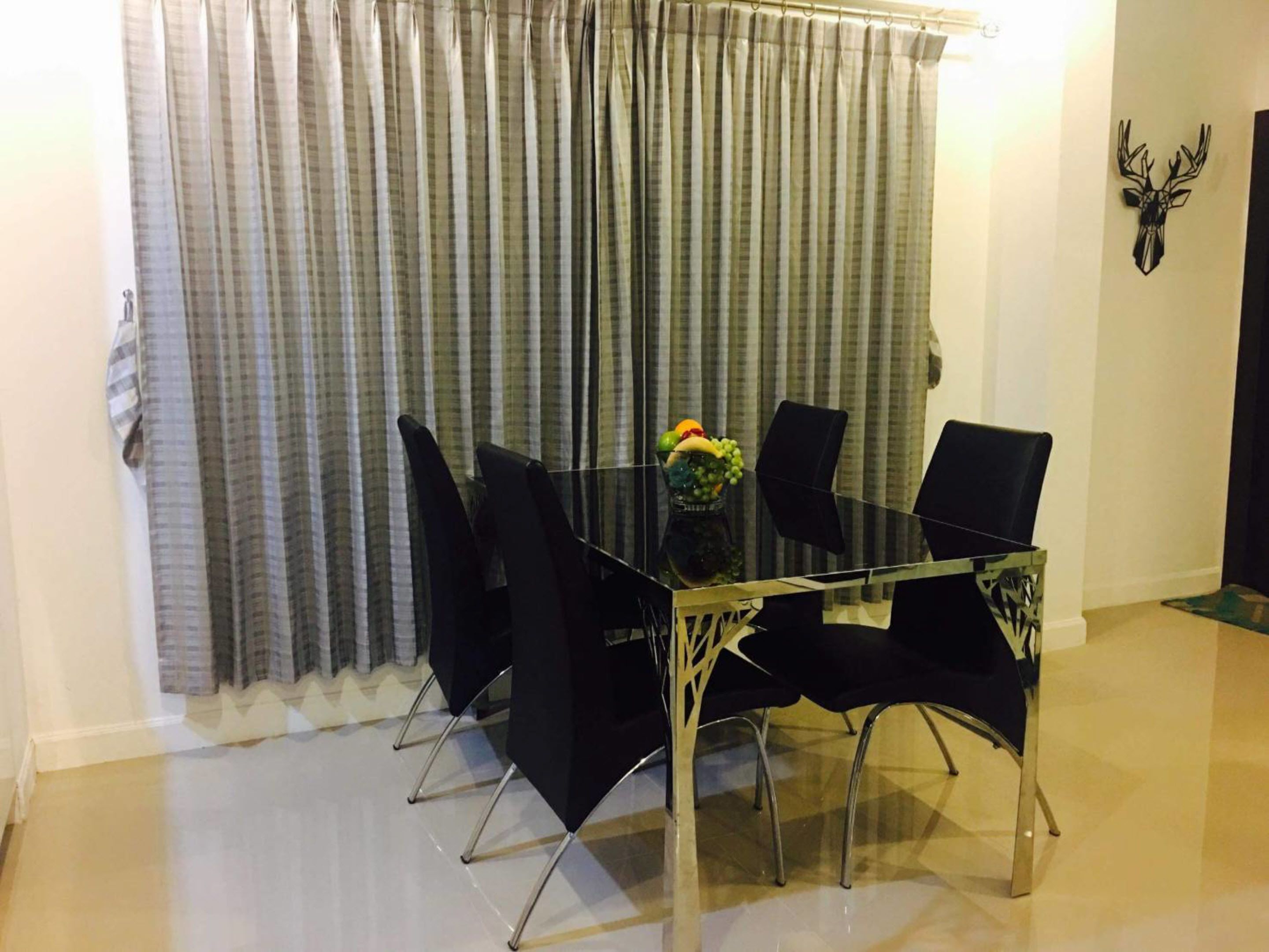 Uma área de refeições com mesa de vidro e cadeiras pretas, um vaso com frutas e uma cortina ao fundo.