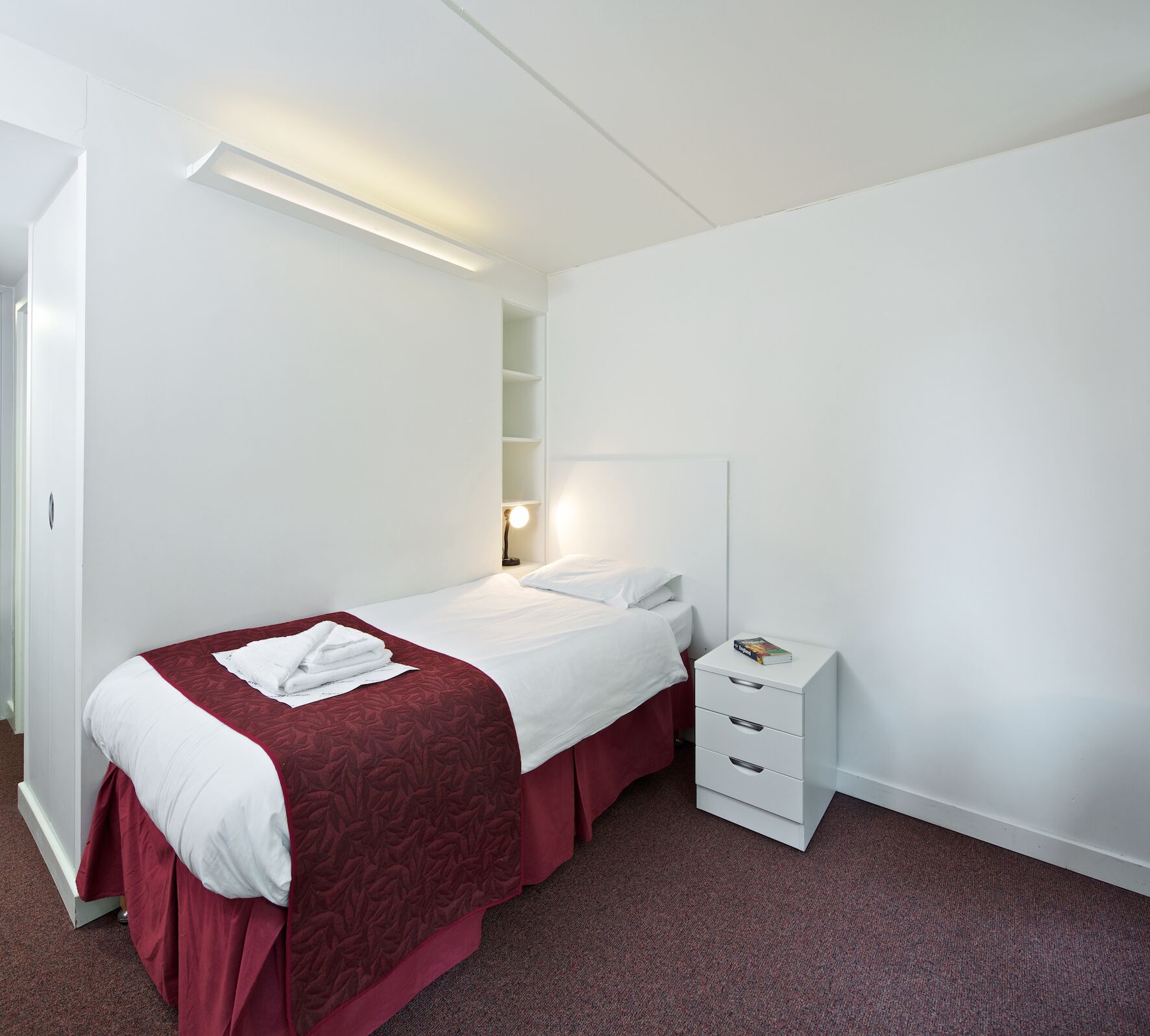 A neatly arranged hotel room with a single bed, a bedside table with drawers, a wall-mounted shelf, and a door.