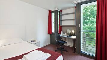 A hotel room with a bed, a desk with a chair, a window with red curtains, and a view of greenery outside.